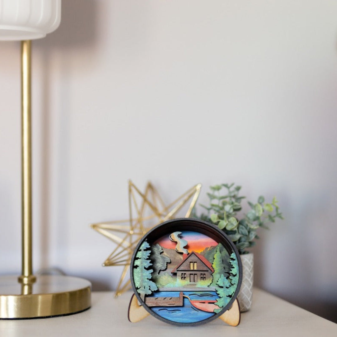 A round decorative ceramic clock with a colorful lakeside cabin scene, displayed on a tabletop next to a gold lamp base, a star-shaped decor object, and a small potted plant.