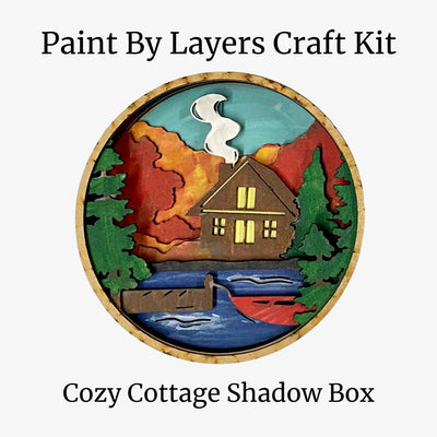 Cozy Cottage Shadow Box craft kit with a cabin, trees, and mountains on a white background.