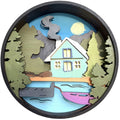 A circular wooden layered art piece depicting a lakeside house scene with pine trees, mountains, a boat, and a yellow sun, shown on a plain white background.