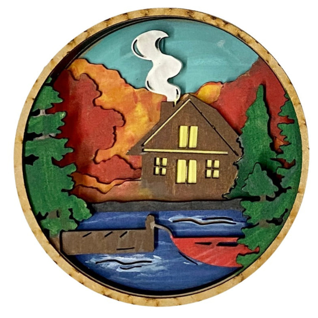 A round wooden plaque featuring a handcrafted, layered landscape design with a brown cabin, red and orange autumn mountains, green pine trees, a blue lake, and a red boat, all framed by a natural cork border.