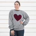 A person wearing a grey crewneck sweater with a red maroon heart stitched to the front of the sweater.
