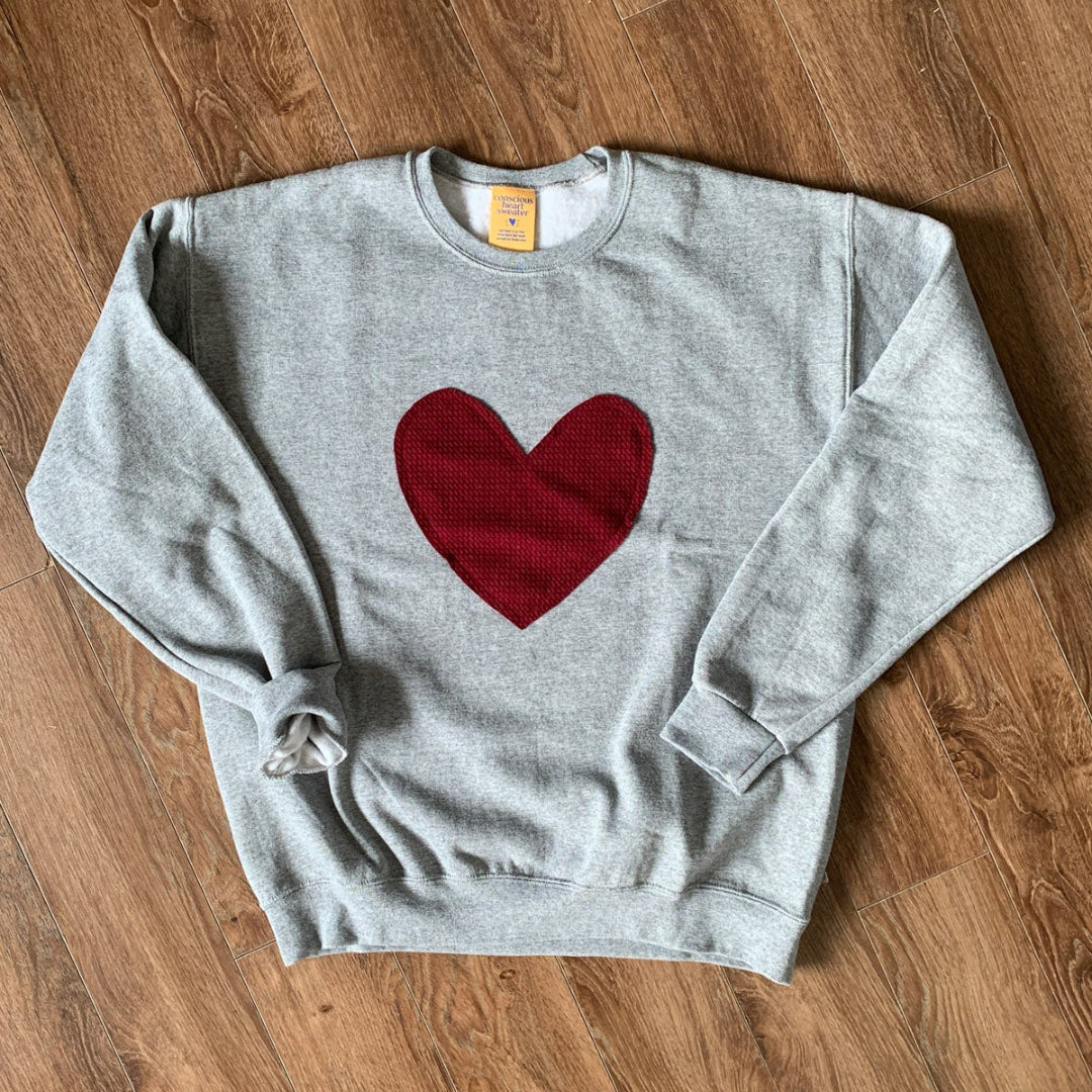 A grey crewneck sweater with a red maroon heart stitched to the front of the sweater.