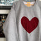 A grey crewneck sweater with a red maroon heart stitched to the front of the sweater.