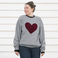 A person wearing a grey crewneck sweater with a red maroon heart stitched to the front of the sweater.
