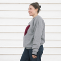 A person wearing a grey crewneck sweater with a red maroon heart stitched to the front of the sweater.