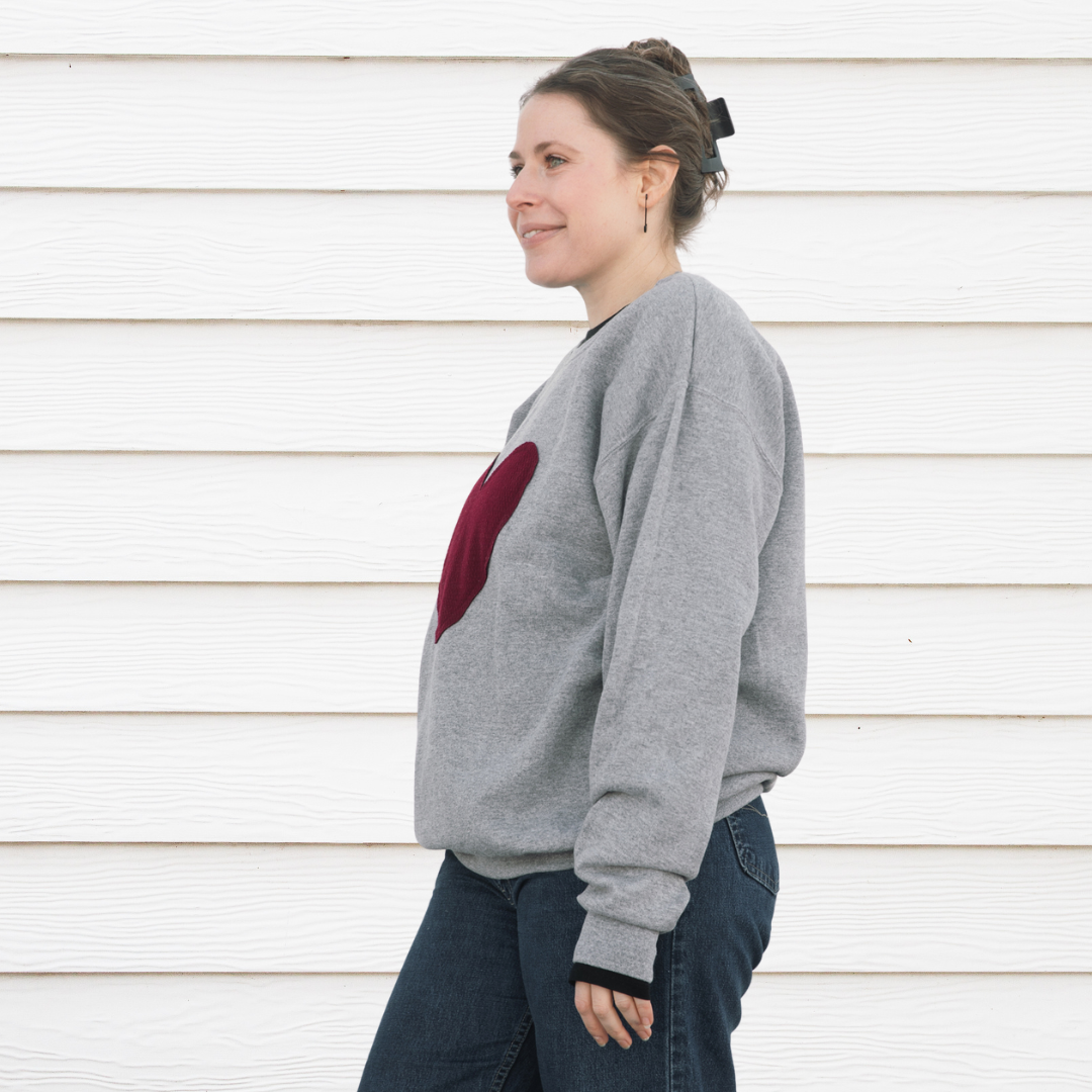 A person wearing a grey crewneck sweater with a red maroon heart stitched to the front of the sweater.