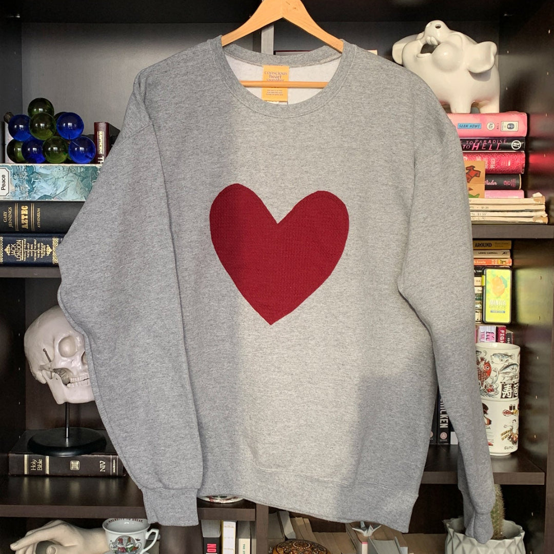 A grey crewneck sweater with a red maroon heart stitched to the front of the sweater.