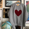 A grey crewneck sweater with a red maroon heart stitched to the front of the sweater.