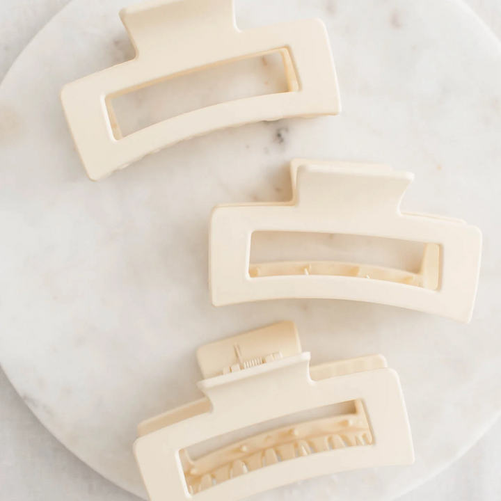 Three large, off-white hair claw clips arranged on a white marble surface. The clips have a smooth, matte finish and a rectangular shape with rounded corners.