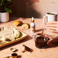 Person pouring a liquid from a dropper into a glass of coffee on a table with food and a bottle.