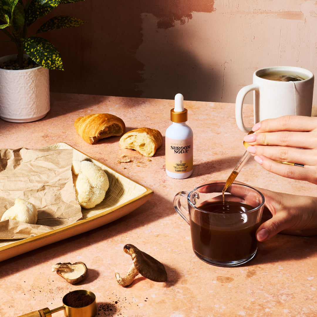 Person pouring a liquid from a dropper into a glass of coffee on a table with food and a bottle.
