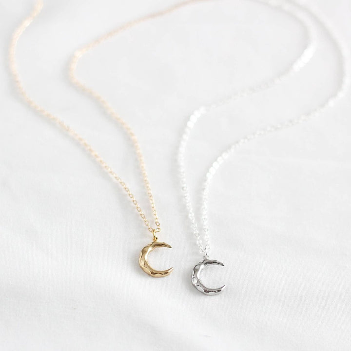 Two crescent moon necklaces on a white cloth. One necklace has a gold-colored crescent moon pendant, and the other has a silver-colored crescent moon pendant. Both necklaces have delicate chains.
