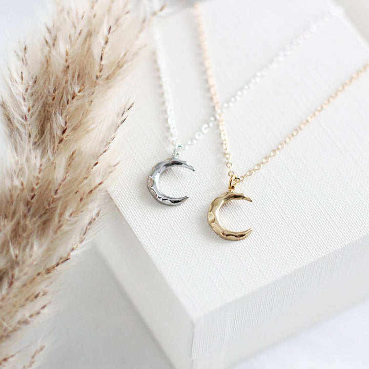 Two crescent moon necklaces on a white cloth. One necklace has a gold-colored crescent moon pendant, and the other has a silver-colored crescent moon pendant. Both necklaces have delicate chains.