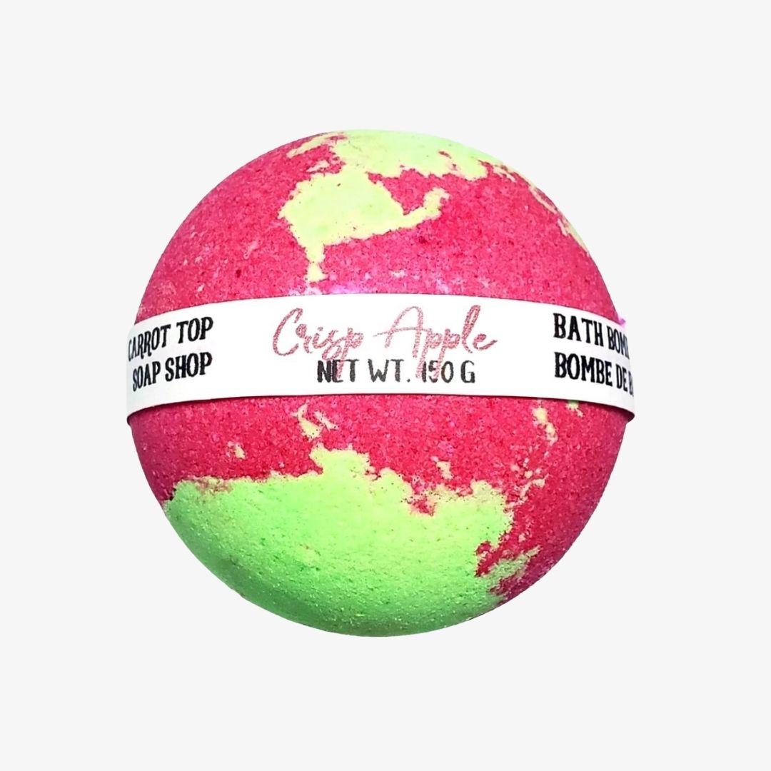 A spherical bath bomb with a pink and neon green marbled pattern, wrapped in a white paper band labeled 'Crisp Apple' and 'Carrot Top Soap Shop'.