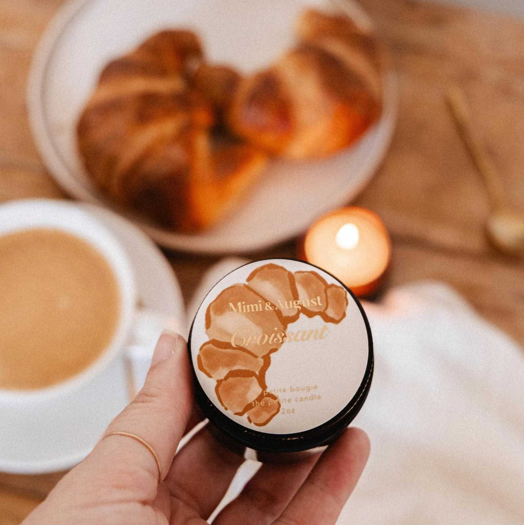 A hand holds a small round tin of Mimic Nature 'amantus' scented candle. The background is a warm, cozy tabletop scene with a plate of croissants, a cup of coffee, and a lit candle.