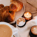 A cozy tabletop scene with croissants on a plate, a cup of coffee, a lit amber candle, and two small round candles with latte-style printed designs on a cream cloth over a wooden table.