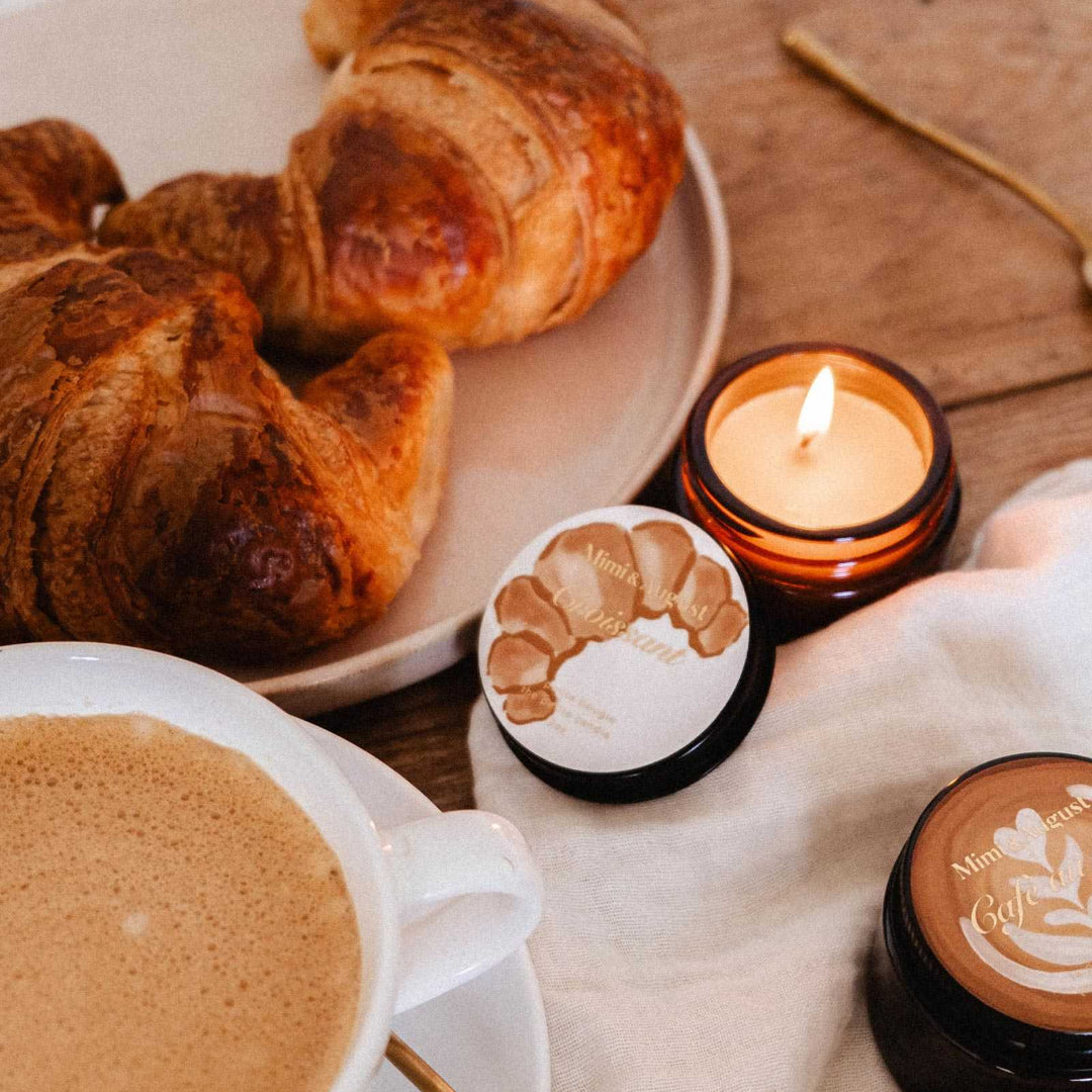 A cozy tabletop scene with croissants on a plate, a cup of coffee, a lit amber candle, and two small round candles with latte-style printed designs on a cream cloth over a wooden table.