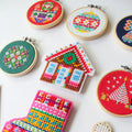 A small, circular embroidery hoop with a festive Christmas scene. The scene depicts a fireplace with candles, presents, and a Christmas tree. The embroidery is colorful and detailed, with a red background.