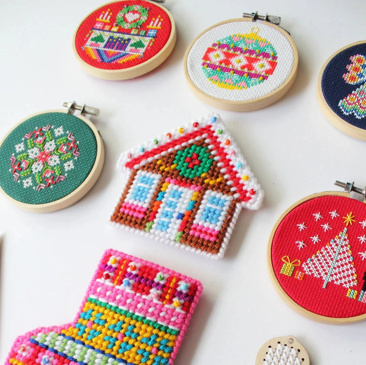 A small, circular embroidery hoop with a festive Christmas scene. The scene depicts a fireplace with candles, presents, and a Christmas tree. The embroidery is colorful and detailed, with a red background.