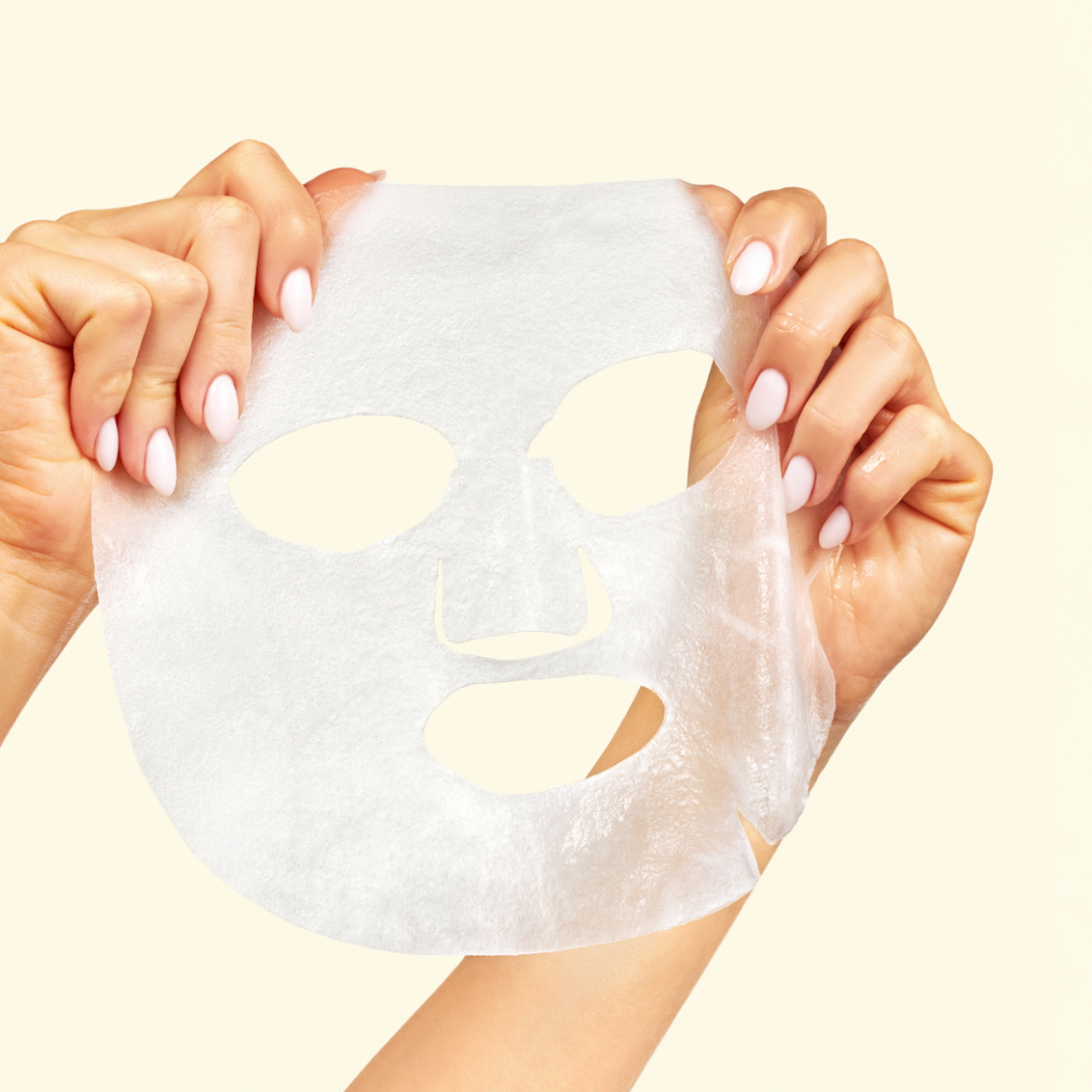 A person holds a translucent white sheet mask against a plain light beige background. The person's hands with pale pink manicured nails are visible on both sides of the mask.