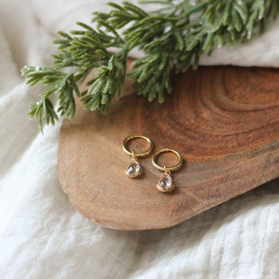 Gold earrings with teardrop-shaped stones on a wooden surface with greenery