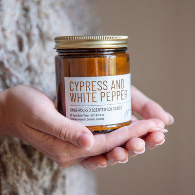 Hand holding a candle labeled 'Cypress and White Pepper' against a neutral background