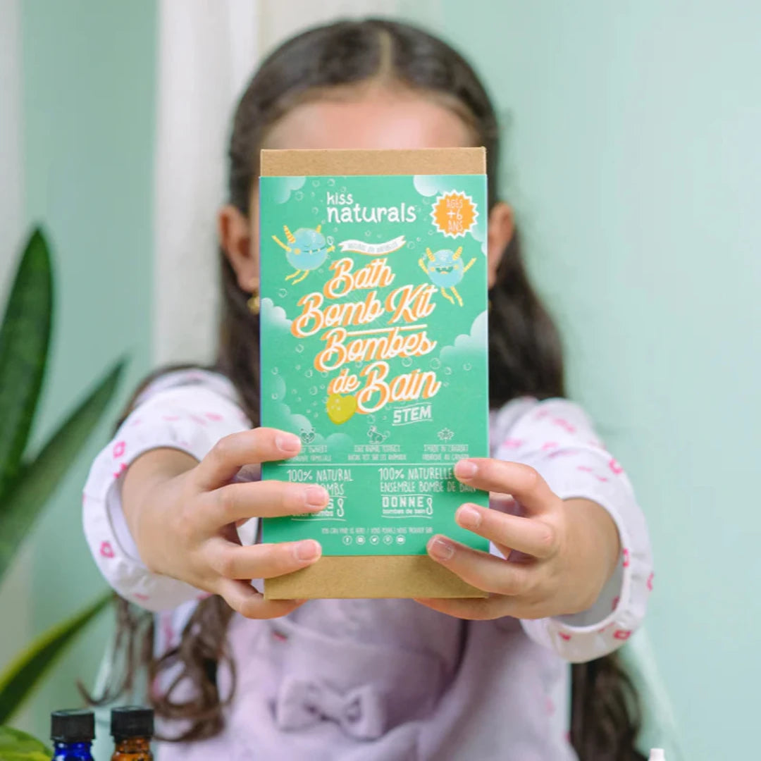 A cardboard box containing a Kiss Naturals Bath Bomb Kit. The box is green and white with the product name and a colorful illustration of bath bombs. It states that the kit is 100% natural, made in Canada, and suitable for ages 6 and up.