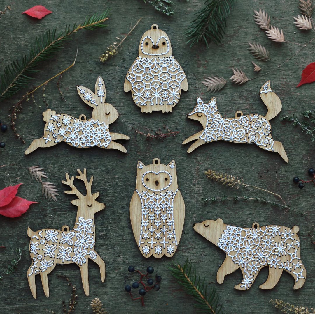A collection of embroidered wooden ornaments depicting various animals, including a rabbit, a penguin, a fox, a deer, an owl, and a bear. The ornaments are decorated with snowflake patterns and are arranged on a green surface with pine needles and red leaves.