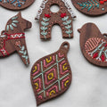 Five embroidered wooden ornaments arranged on a white surface. The ornaments depict a snowflake, a gingerbread house, a Dala horse, a bird, and a geometric design. The ornaments are decorated with colorful embroidery stitches.