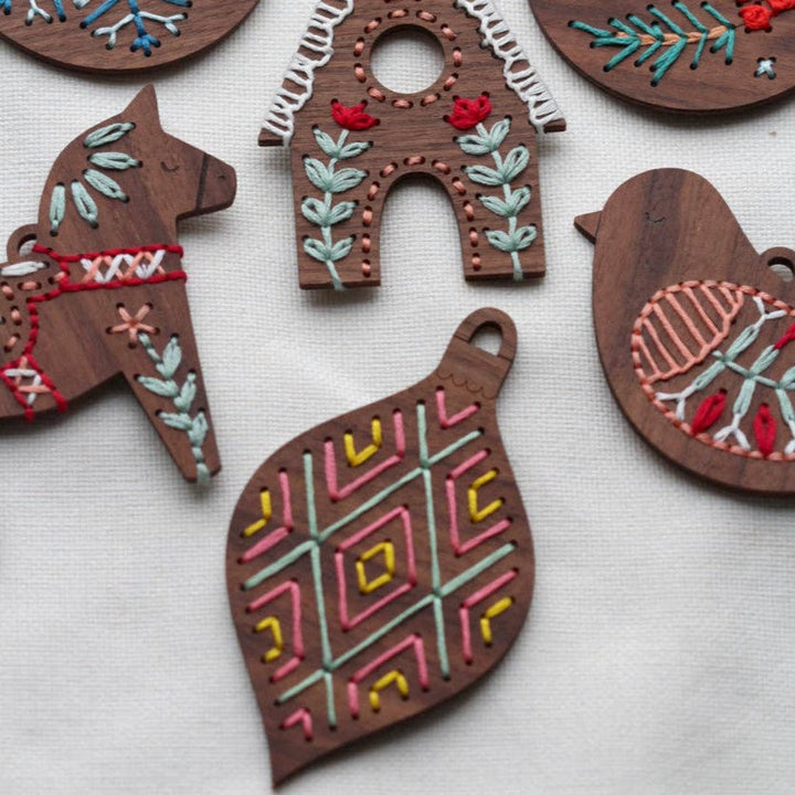 Five embroidered wooden ornaments arranged on a white surface. The ornaments depict a snowflake, a gingerbread house, a Dala horse, a bird, and a geometric design. The ornaments are decorated with colorful embroidery stitches.