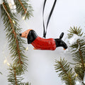 A glossy ceramic dachshund Christmas ornament wearing a red sweater with black spots, hanging from a black ribbon against a white background with pine branches and fairy lights.