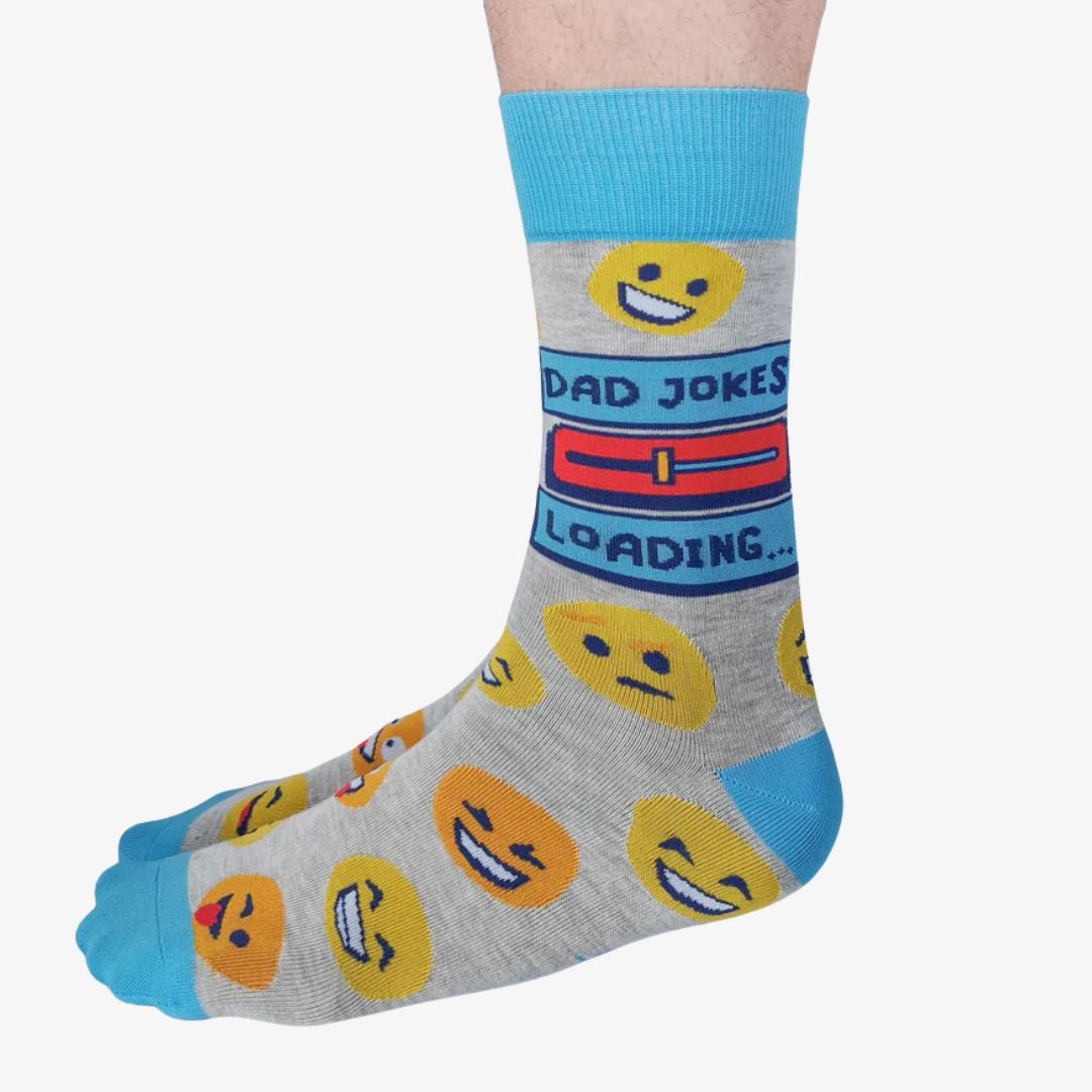 A pair of grey crew socks featuring a "Dad Jokes Loading..." graphic with a progress bar in red and blue, surrounded by various yellow and orange emoji faces. The text "DAD JOKES" is written in blue above the loading bar. The cuff, heel, and toe of the sock are light blue.