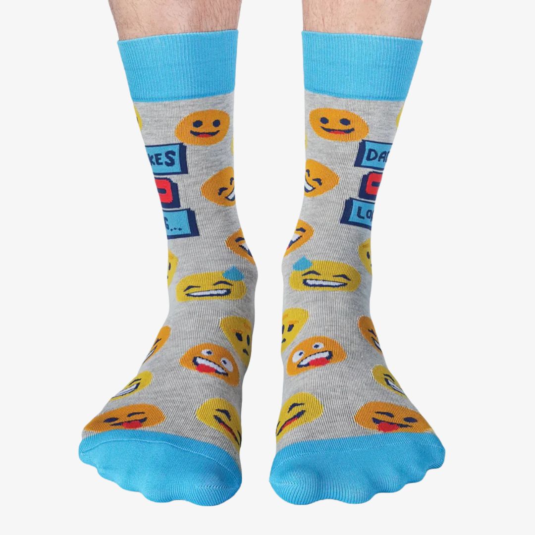 A pair of grey crew socks featuring a "Dad Jokes Loading..." graphic with a progress bar in red and blue, surrounded by various yellow and orange emoji faces. The text "DAD JOKES" is written in blue above the loading bar. The cuff, heel, and toe of the sock are light blue.