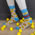 A pair of grey crew socks featuring a "Dad Jokes Loading..." graphic with a progress bar in red and blue, surrounded by various yellow and orange emoji faces. The text "DAD JOKES" is written in blue above the loading bar. The cuff, heel, and toe of the sock are light blue.