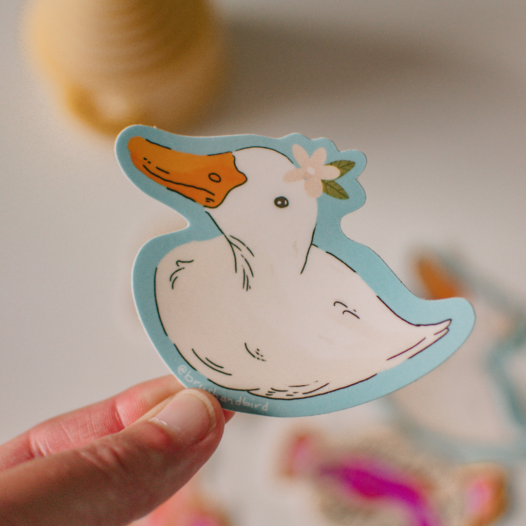 Hand holding a sticker of a cartoon duck with a flower on its head against a blurred background.