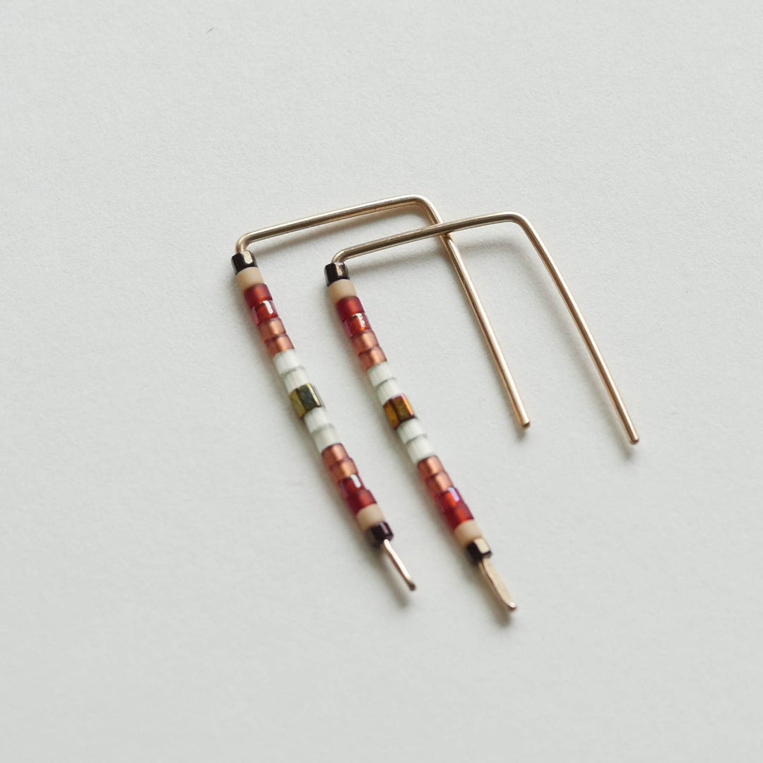 A pair of minimalist U-shaped stud earrings crafted from delicate 14 KT gold wire. Each earring features a short segment of small Japanese glass seed beads in a pattern of red, beige, white, olive green, and brown near the top of the hanging bar.