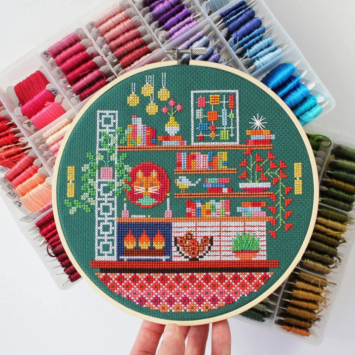 A completed cross-stitch project in a wooden hoop. The design depicts a cozy living room scene with a fireplace, bookshelves, plants, and a cat. The scene is filled with colorful details and is set against a green background. The text 