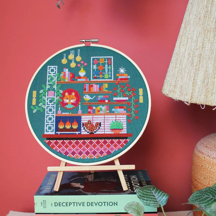 A completed cross-stitch project in a wooden hoop. The design depicts a cozy living room scene with a fireplace, bookshelves, plants, and a cat. The scene is filled with colorful details and is set against a green background. The text 