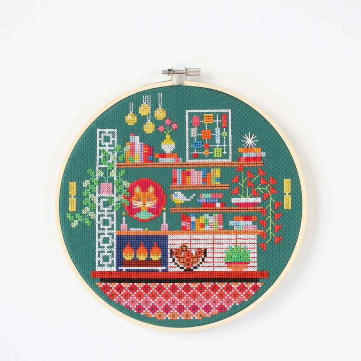 A completed cross-stitch project in a wooden hoop. The design depicts a cozy living room scene with a fireplace, bookshelves, plants, and a cat. The scene is filled with colorful details and is set against a green background. The text 