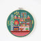 A completed cross-stitch project in a wooden hoop. The design depicts a cozy living room scene with a fireplace, bookshelves, plants, and a cat. The scene is filled with colorful details and is set against a green background. The text 