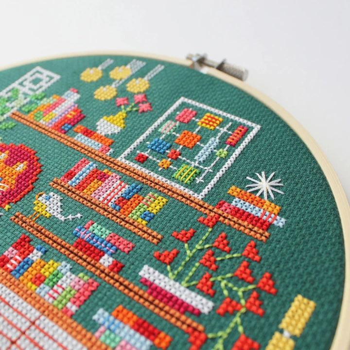 A completed cross-stitch project in a wooden hoop. The design depicts a cozy living room scene with a fireplace, bookshelves, plants, and a cat. The scene is filled with colorful details and is set against a green background. The text 