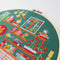 A completed cross-stitch project in a wooden hoop. The design depicts a cozy living room scene with a fireplace, bookshelves, plants, and a cat. The scene is filled with colorful details and is set against a green background. The text 