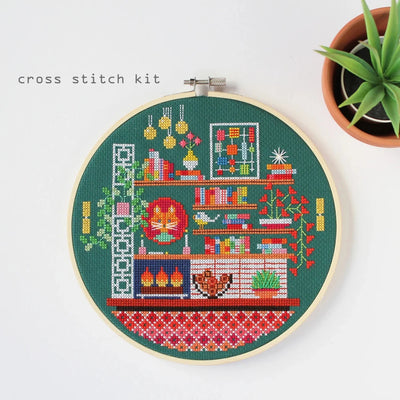 A completed cross-stitch project in a wooden hoop. The design depicts a cozy living room scene with a fireplace, bookshelves, plants, and a cat. The scene is filled with colorful details and is set against a green background. The text 
