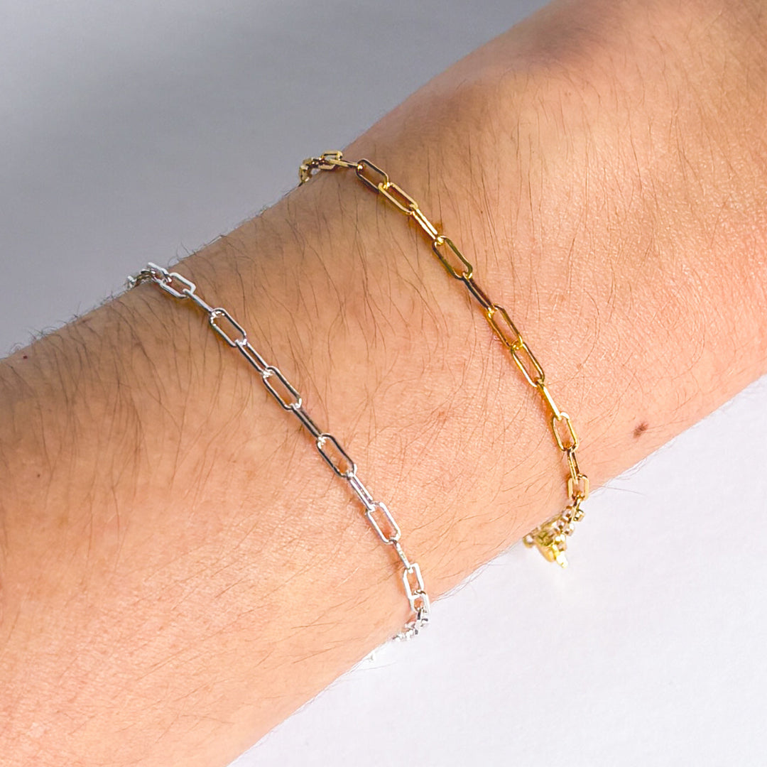 Two chain bracelets, one gold and one silver, on a wrist against a neutral background.