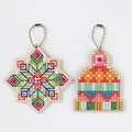 Two wooden cross-stitch ornaments on a white background, one shaped like a snowflake with a geometric multicolor pattern and the other like a knit hat with a colorful geometric pattern. Both have silver ball chains.