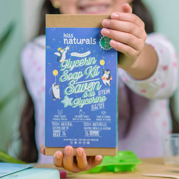 A cardboard box containing a Kiss Naturals Glycerin Soap Kit. The box is blue and white with the product name and a colorful illustration of penguins. It states that the kit is 100% natural, made in Canada, and suitable for ages 6 and up.