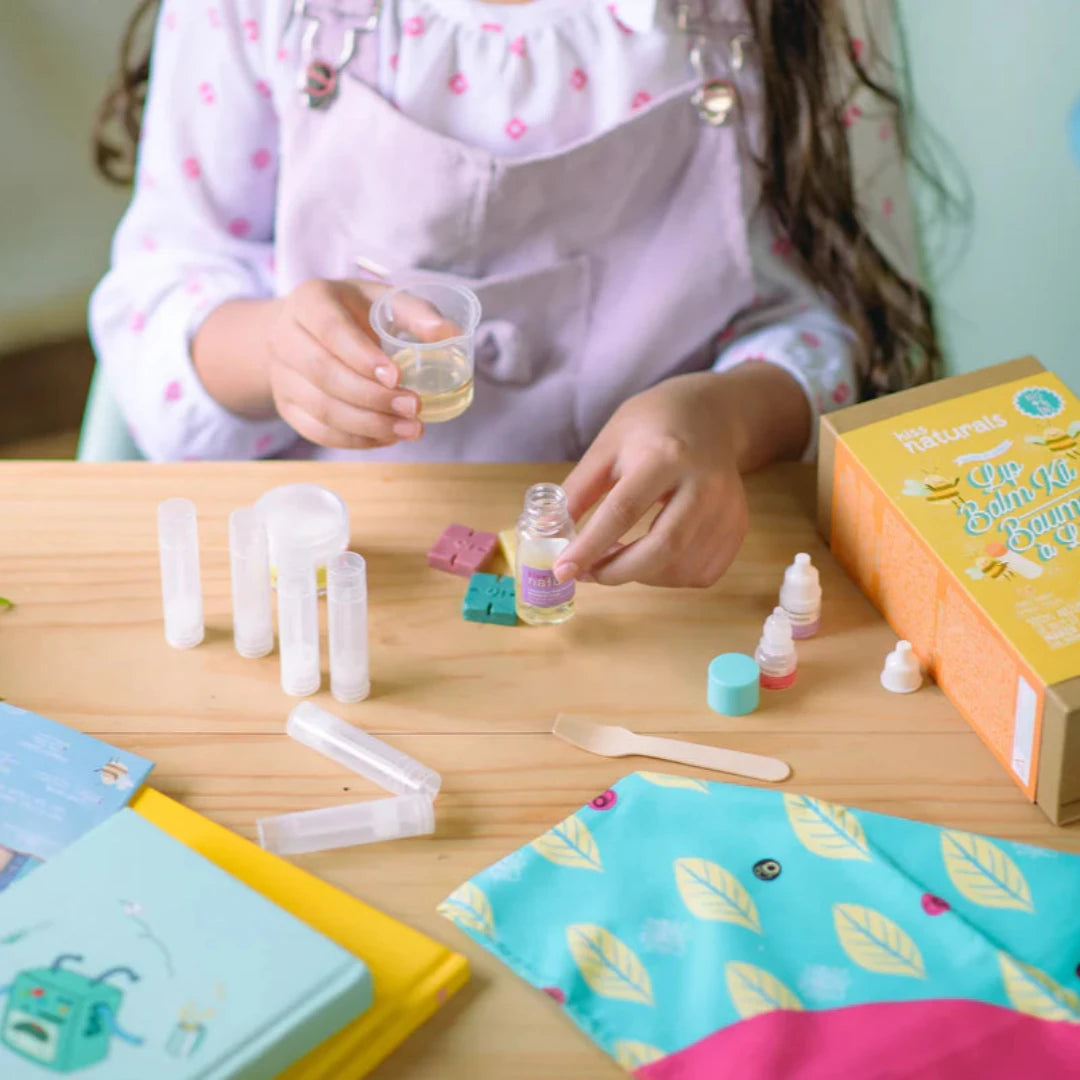 A cardboard box containing a Kiss Naturals Lip Balm Kit. The box is yellow and white with the product name and a colorful illustration of bees. It states that the kit is 100% natural, made in Canada, and suitable for ages 6 and up.