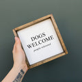 Hand holding a framed sign with 'DOGS WELCOME people tolerated' text on a dark background