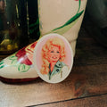 An oval pin featuring a vintage-style illustration of Dolly Parton, displayed on a rustic wooden surface next to hand-painted cowboy boots.