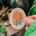 A hand holds an oval sticker featuring a retro-style illustration of a woman with blonde hair and a green shirt with a shamrock, set against a cozy, dark background with plants and a rabbit figurine.